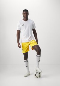 Camiseta blanca de manga corta con el logo negro de Adidas, pantalones cortos de fútbol amarillos, calcetines blancos con rayas negras y botas de fútbol blancas, sosteniendo un balón de fútbol.