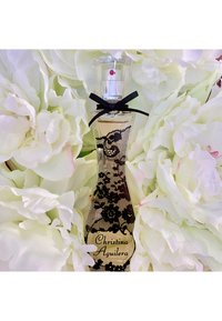Flacon de parfum avec un motif en dentelle noire et un nœud noir, étiqueté « Christina Aguilera », entouré de grandes fleurs blanches.