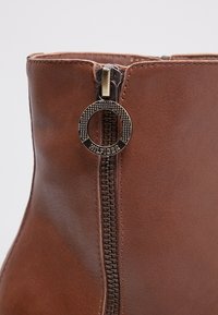 Bottine en cuir marron avec une fermeture éclair en métal visible et un tirant circulaire, gravé du nom de la marque. Texture lisse sur toute la surface.