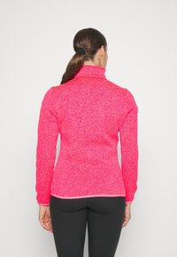 Kobieta z ciemnymi włosami związanymi w kucyk, ubrana w jasnoróżową długą bluzę i czarne legginsy, stojąca przodem do białej ściany, z widocznymi plecami.