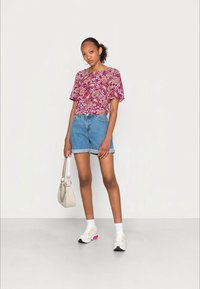 Blusa bordada em padrão floral maroon, calções de ganga claros, sapatilhas brancas e rosas, e uma pequena mala bege. Modelo está de pé contra um fundo neutro.