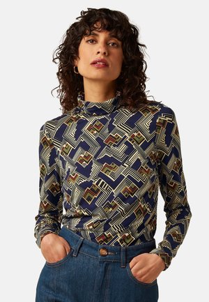 Vrouw met krullend haar die een top met lange mouwen en een hoge hals draagt met een geometrisch patroon in marineblauw, olijfgroen en beige, en een blauwe spijkerbroek met handen in de zakken.
