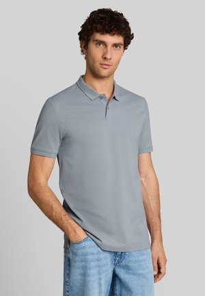 Man met krullend haar die een lichtgrijs poloshirt met korte mouwen en lichtblauwe spijkerbroek draagt, staat met één hand in de zak.