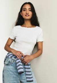 Weißes, geripptes Oberteil mit gewelltem Ausschnitt und Ärmelsaum, kombiniert mit hellblau-navy gestreiftem Stoff, der in der Taille gebunden ist, getragen zu Denim-Jeans.