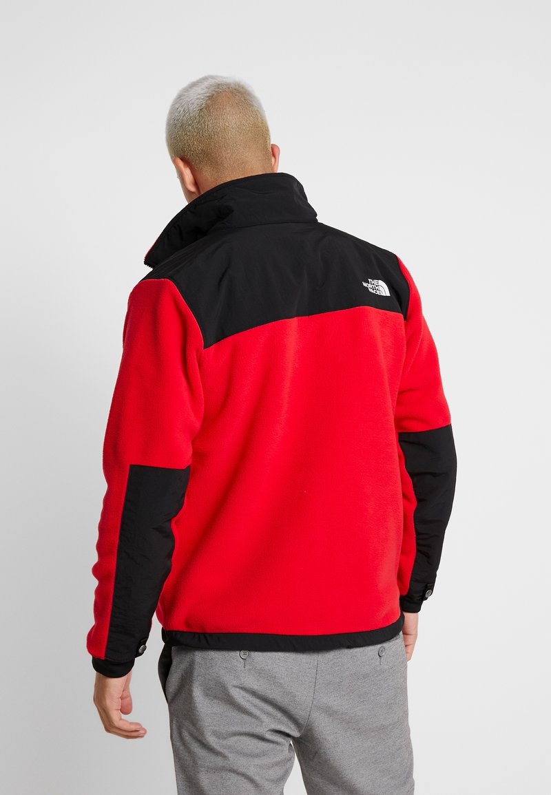 Uomo con capelli biondi corti che indossa una giacca in pile rossa e nera con il logo The North Face, in piedi contro uno sfondo grigio uniforme.