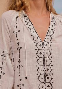 Hvid bluse med detaljeret sort broderi omkring halsen og ærmerne, med en V-formet åbning og snøre lukning.