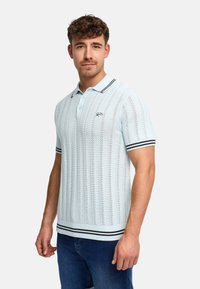 Uomo che indossa una polo lavorata a maglia azzurro chiaro con colletto e bordi delle maniche a righe bianche e nere, abbinata a jeans blu, in piedi su sfondo bianco.