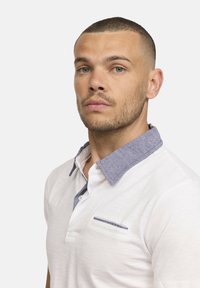 Polo blanc avec un col en chambray bleu contrastant et une petite poche avec une rayure bleue sur la poitrine. Texture lisse, design décontracté.