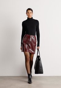Schwarzes Rollkragenoberteil, glänzender bordeauxroter Rock mit hoher Taille, kombiniert mit schwarzen Knöchel-Stiefeln und einer großen schwarzen Handtasche. Glatte Texturen überall.