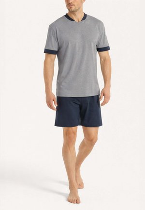 Uomo che cammina a piedi nudi, indossando una maglietta grigia a maniche corte con rifiniture blu navy e pantaloncini blu navy su uno sfondo semplice.