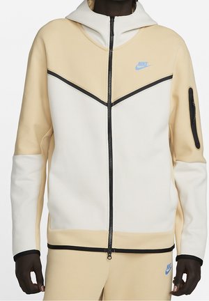 Person trägt beige-weiße zweifarbige Nike Kapuzenjacke mit Reißverschluss, schwarzer Einfassung und kleinen blauen Nike-Logos auf Brust und Hose.