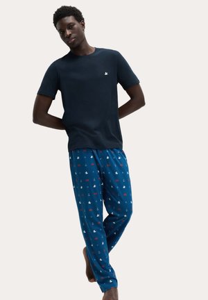 Młody mężczyzna ubrany w granatowy t-shirt i niebieskie spodnie do spania z białymi, czerwonymi i granatowymi nadrukami o tematyce morskiej, stojący boso z rękami za plecami.
