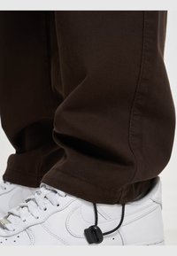 Pantalones marrones ajustados con una textura suave, que presentan una costura recta y un leve dobladillo en el bajo, combinados con zapatillas blancas con cordones.