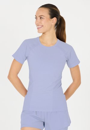 Femme avec une queue de cheval brune portant un haut à manches courtes violet clair et un short assorti, souriante et regardant sur le côté les mains derrière le dos.