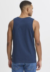 Tumši zils bezdelņu tops, kas izgatavots no mīksta kokvilnas, ar apaļu kakla izgriezumu un brīvu piegriezumu, pie apakšmalas redzama birka. Valkāts ar džinsa šortiem.