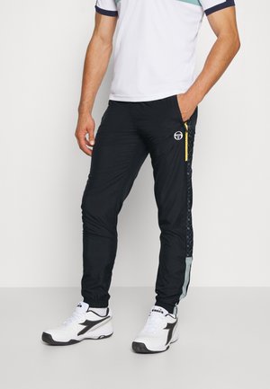 Mann trägt schwarze Sporthose mit gelbem Reißverschlussdetail und weiße Sneaker mit schwarzen Akzenten, steht vor einem schlichten Hintergrund.