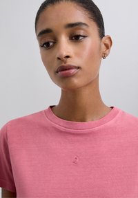 T-shirt en coton rose avec un col rond et des manches courtes. Présente un petit logo en relief au centre de la poitrine. Texture lisse.