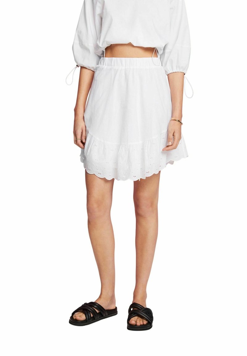 Gonna bianca in cotone con orlo arricciato, caratterizzata da bordo scallop e dettagli in eyelet. Abbinata a sandali neri a scivolo. Stile casual, vestibilità comoda e ampia.