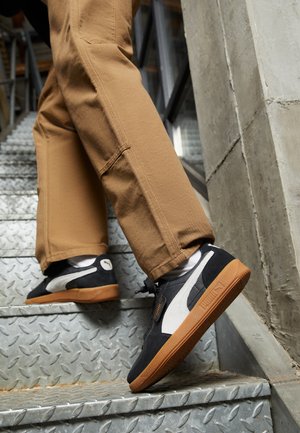 Zapatillas de ante negras y grises con detalles blancos y suelas de goma, llevadas con pantalones beige mientras se suben escaleras de metal.