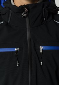 Svart jacka med huva, med blå dragkedjedetaljer, två framfickor med dragkedja och en glansig EA-logotyp på kragen. Strukturerad tyg.