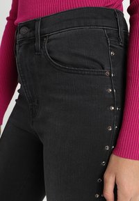 Primer plano de una persona que lleva unos jeans negros con tachuelas metálicas a lo largo de la costura lateral y una camiseta de manga larga de magenta con relieve.