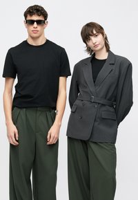 Μαύρο βαμβακερό T-shirt, πράσινο πλισέ παντελόνι. Γκρι σακάκι διπλό πετσετέ με ζώνη. Το μοντέλο φοράει γυαλιά ηλίου και σκουλαρίκια.