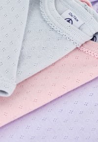 Tre strikkede bomullsklær i lys blå, rosa og lavendel, med et strukturert mønster med små hull og bølgete halslinjer.