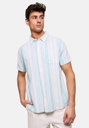 Hombre con camisa de manga corta abotonada con rayas verticales blancas y azul claro, posando con una mano en el bolsillo, mirando de lado.