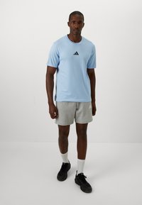 Lyseblå treningst-skjorte, kortermet, svart logo på brystet, kombinert med grå shorts, svarte joggesko og hvite sokker. Enkelt design.