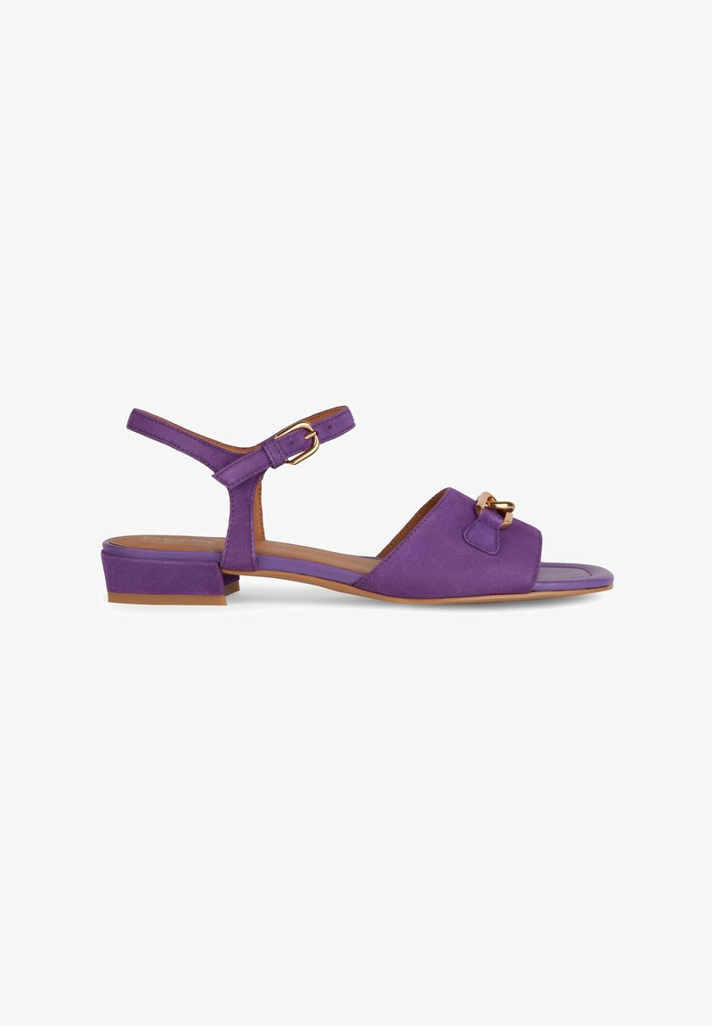 Sandale en daim violet avec un petit talon carré, bride de cheville ajustable et détail décoratif en nœud sur l'avant-pied. Semelle plate, design minimaliste.