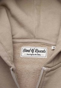 Beige huvtröja med en slät yta och mjukt fleecefoder. Har en dragkedja och en etikett med texten "Band of Rascals." Ekologiskt tyg.