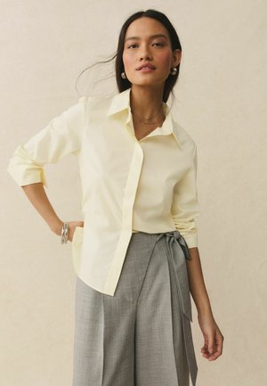 Woman with long dark hair wears a light yellow collared shirt with rolled-up sleeves and high-waisted gray pants with a side tie.