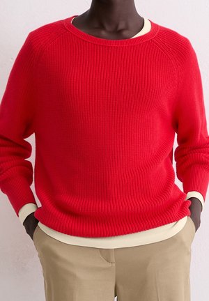 Personne portant un pull rouge vif côtelé superposé à une chemise crème à manches longues avec un pantalon kaki, mains dans les poches.
