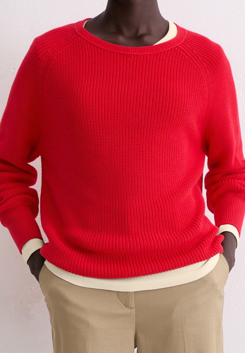 Personne portant un pull rouge vif côtelé superposé à une chemise crème à manches longues avec un pantalon kaki, mains dans les poches.