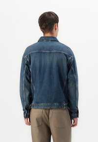 Person wearing a blue denim jacket and beige pants, standing with back facing camera against a plain white background.