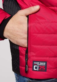 Chaqueta acolchada roja con un panel lateral negro, bolsillo con cremallera y un parche de logo con la inscripción "CAMP DAVID" en azul y blanco.