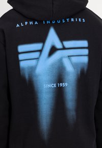 Svart hettegenser med blå spraymalt design av en stilisert «A» og tre horisontale linjer på baksiden, med «SINCE 1959» under.