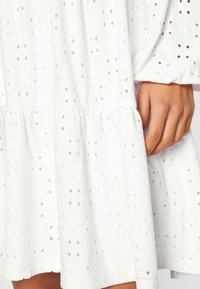 Main reposant contre une robe blanche en tissu à œillets avec un petit motif circulaire de découpe et un détail de couture froncée.