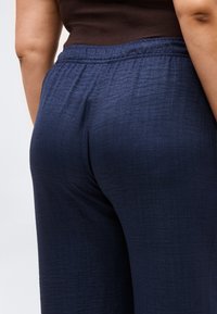 Άτομο που φοράει σκούρο μπλε υφασμάτινο παντελόνι με ελαστική μέση και εφαρμοστή σκούρα καφέ μπλούζα, εμφανιζόμενο από τη μέση και κάτω, από την πίσω όψη.