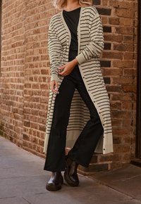 Béžový dlouhý cardigan s černými horizontálními pruhy, lehký pletený materiál, knoflíky, nošený přes černé tričko a kombinovaný s černými kalhotami a kozačkami.