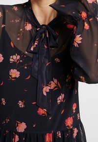 Abito floreale blu navy con maniche lunghe trasparenti, scollo con laccetto e motivo di fiori rosa; presenta un orlo arricciato e tessuto leggero.