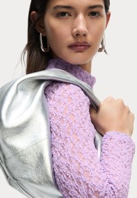 Top in pizzo lavanda con motivi texture, abbinato a una borsa argentata lucida appoggiata sulla spalla. Sfondo neutro.