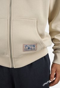 Beige zip-up hette med en frontlomme, ribbestrikkede mansjetter og kant, samt en blå og rød PEGADOR-lapp med teksten "Everyday Ambition".