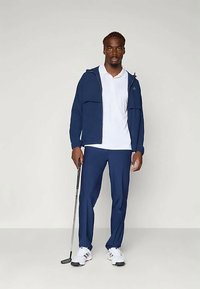 Tamsiai mėlyna užtrauktuku užsegama striukė ant baltos polo marškinėlių, derinama su tamsiai mėlynomis kelnėmis ir baltomis golfo bateliais. Asmuo laiko golfo lazda.