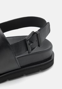 Sandalo in pelle nera con una fascia ampia e fibbia regolabile. Suola spessa con una finitura testurizzata, progettata per resistere e garantire comfort.