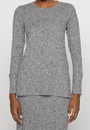 Kobieta ubrana w szary, długorękawowy, teksturowany sweter i pasującą spódnicę, stojąca z ramionami luźno opuszczonymi wzdłuż ciała na jednolitym tle.