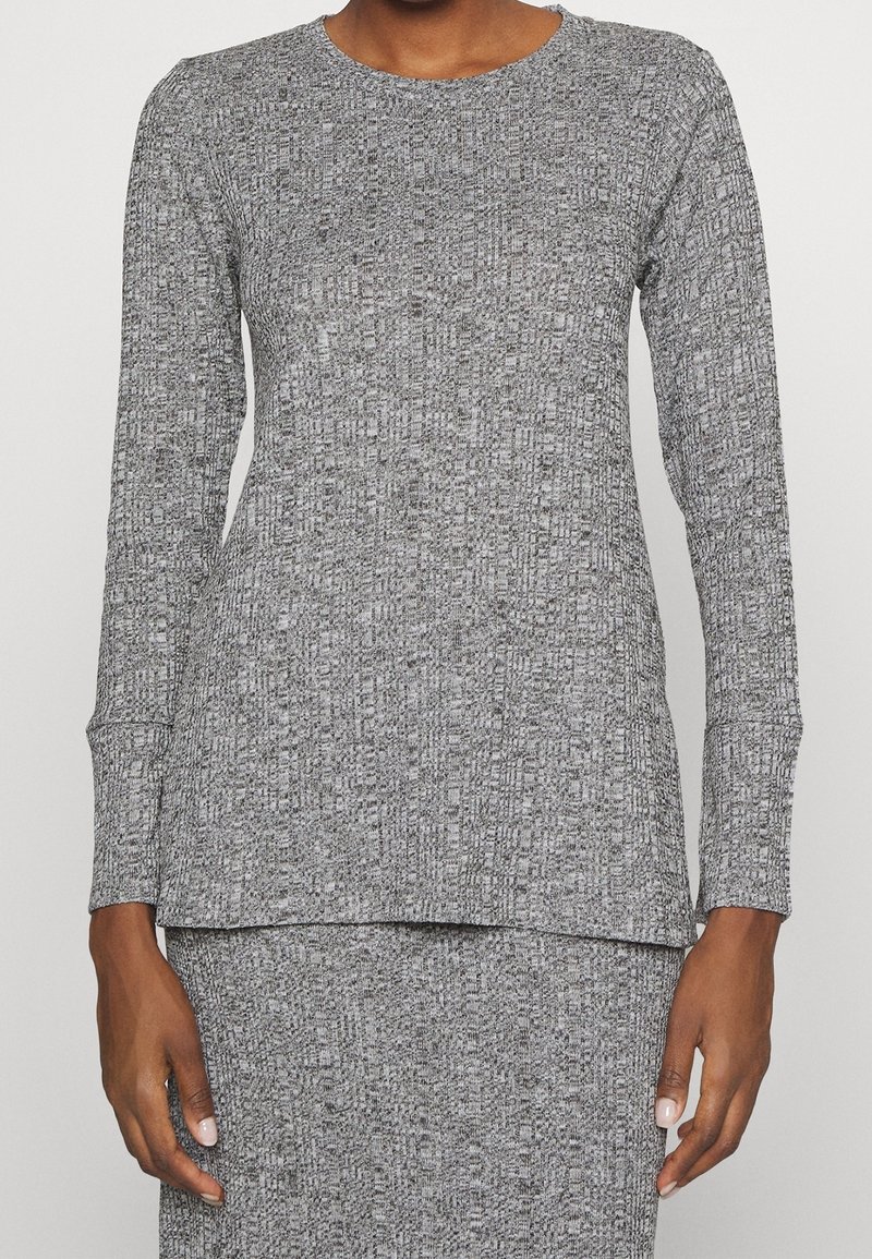 Femme portant un haut à manches longues en maille grise texturée et une jupe assortie, debout avec les bras détendus le long du corps, sur un fond uni.