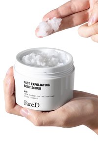 White round container of body scrub with a smooth, glossy finish. Scrub is granular and white, held between fingers. Label includes product name.