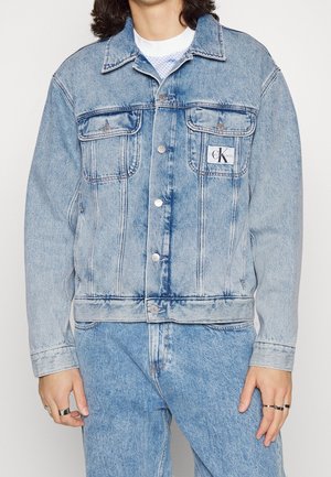 Osoba nosząca jasnoniebieską kurtkę dżinsową Calvin Klein na białej koszuli, w połączeniu z pasującymi dżinsami z wysokim stanem i srebrnymi pierścionkami.