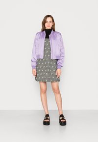 Casaco bomber lilás sobre um vestido xadrez preto e branco com detalhes florais amarelos, combinado com sandálias de plataforma pretas e grossas.